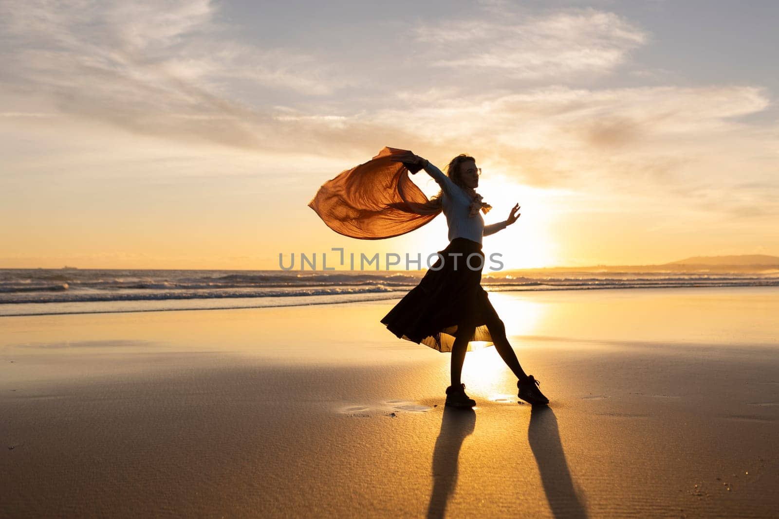 A woman is dancing on the beach with a scarf in her hand. The sky is orange and the sun is setting