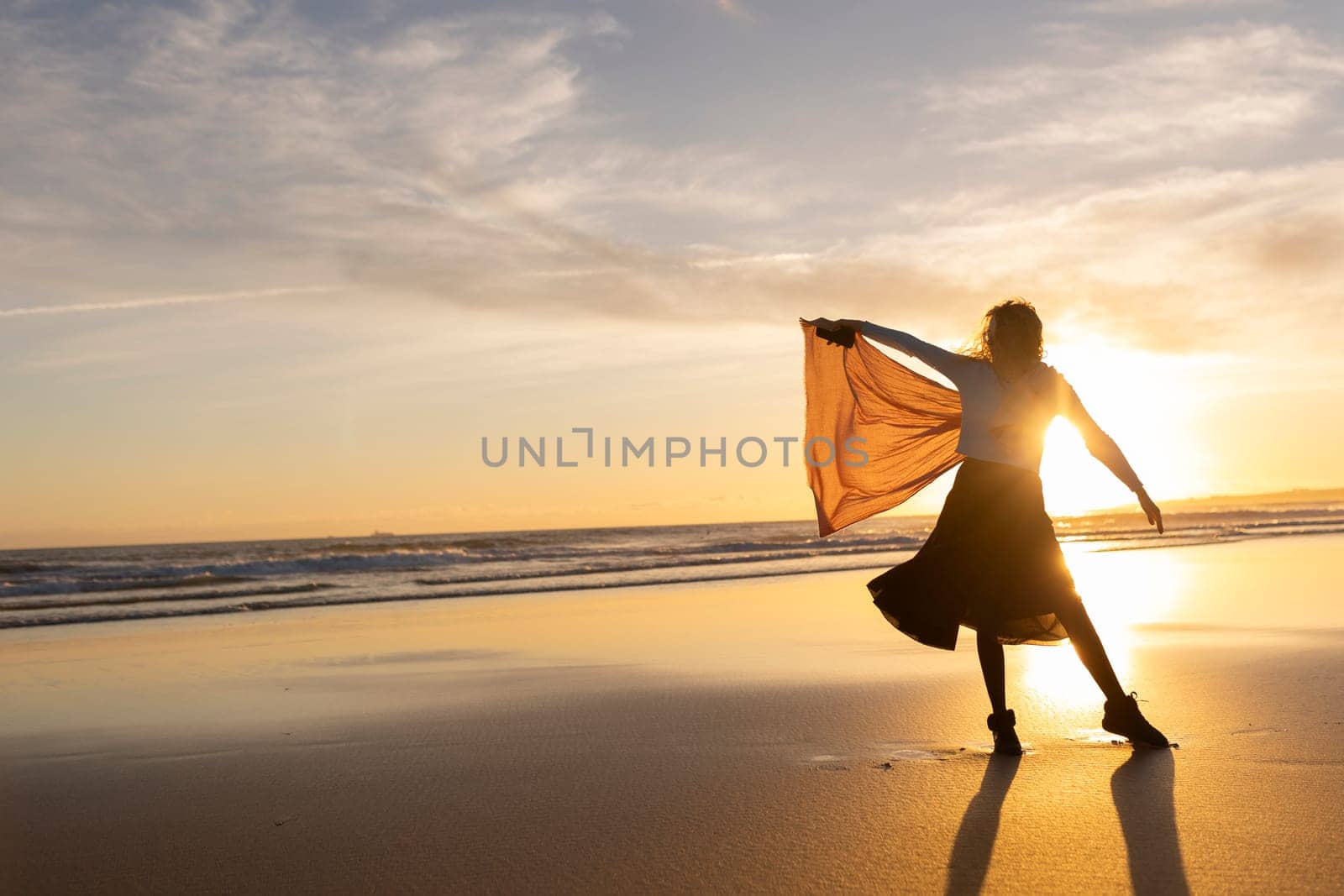 A woman is standing on the beach with a scarf in her hand. She is dancing and enjoying the sunset