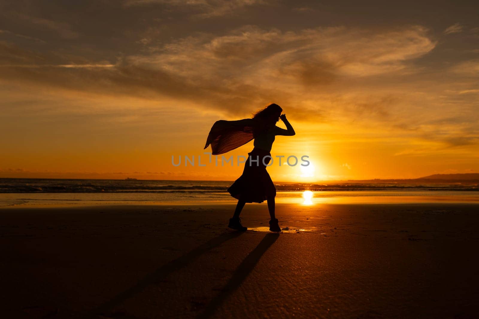 A woman is walking on the beach with a scarf in her hand. The sky is orange and the sun is setting