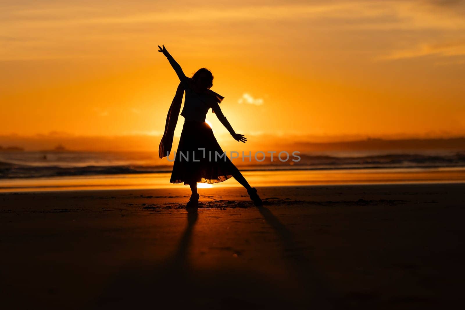 A woman is dancing on the beach at sunset. She is wearing a skirt and a scarf. The scene is peaceful and serene, with the sun setting in the background