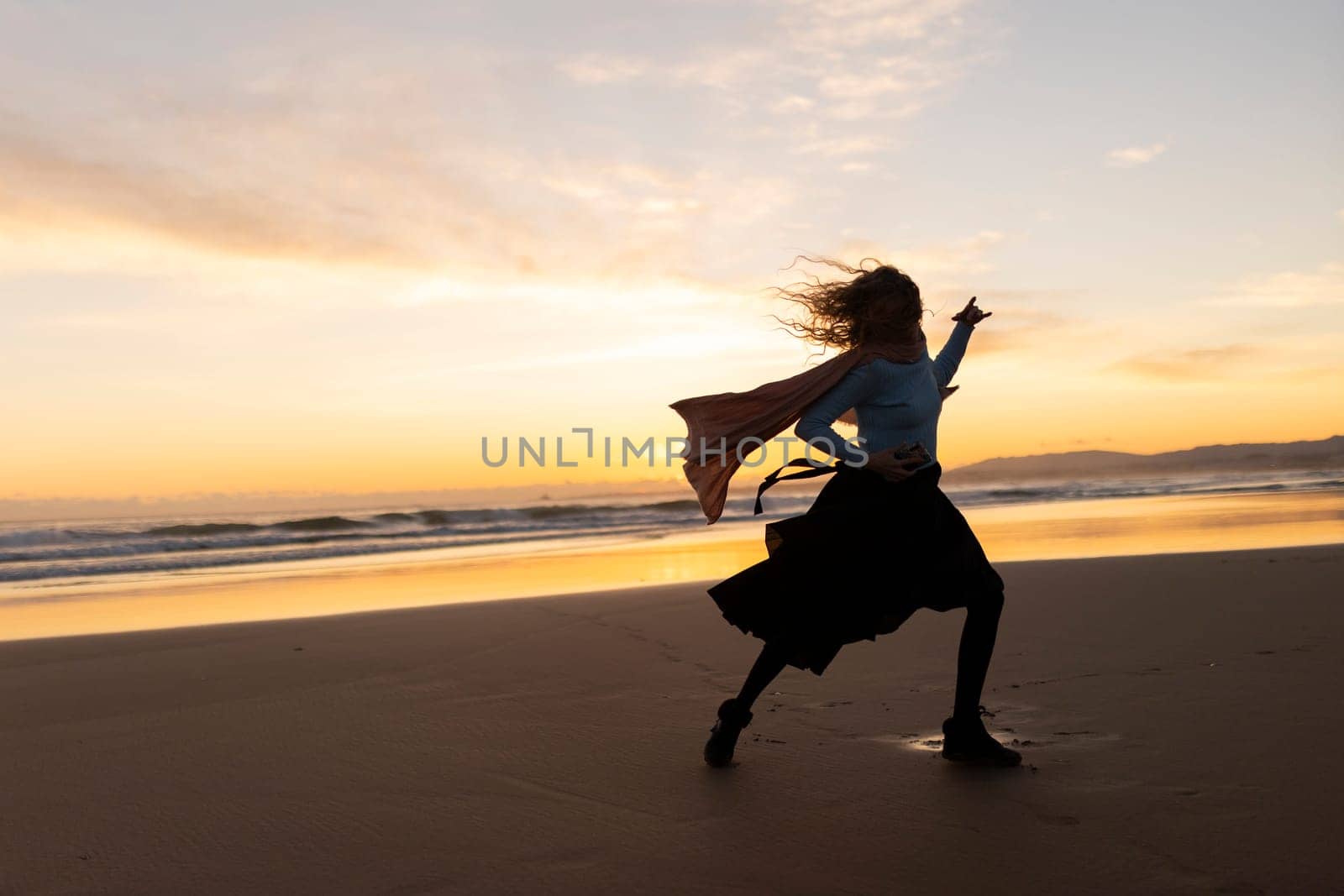 A woman is running on the beach with a scarf around her neck. The sky is orange and the water is calm