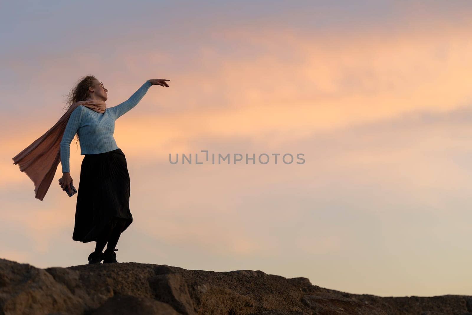 A woman is standing on a hill with a scarf around her neck. She is looking up at the sky, which is filled with clouds. The sky is a mix of orange and pink hues, creating a serene