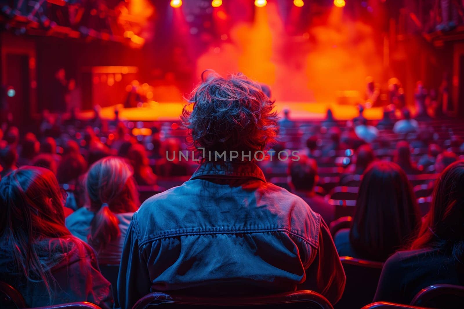 A man sits in the back row of a theater auditorium, watching a performance on stage. The stage is brightly lit with red spotlights, while the audience is bathed in blue light. The image captures the anticipation and excitement of being in the audience at a live show.