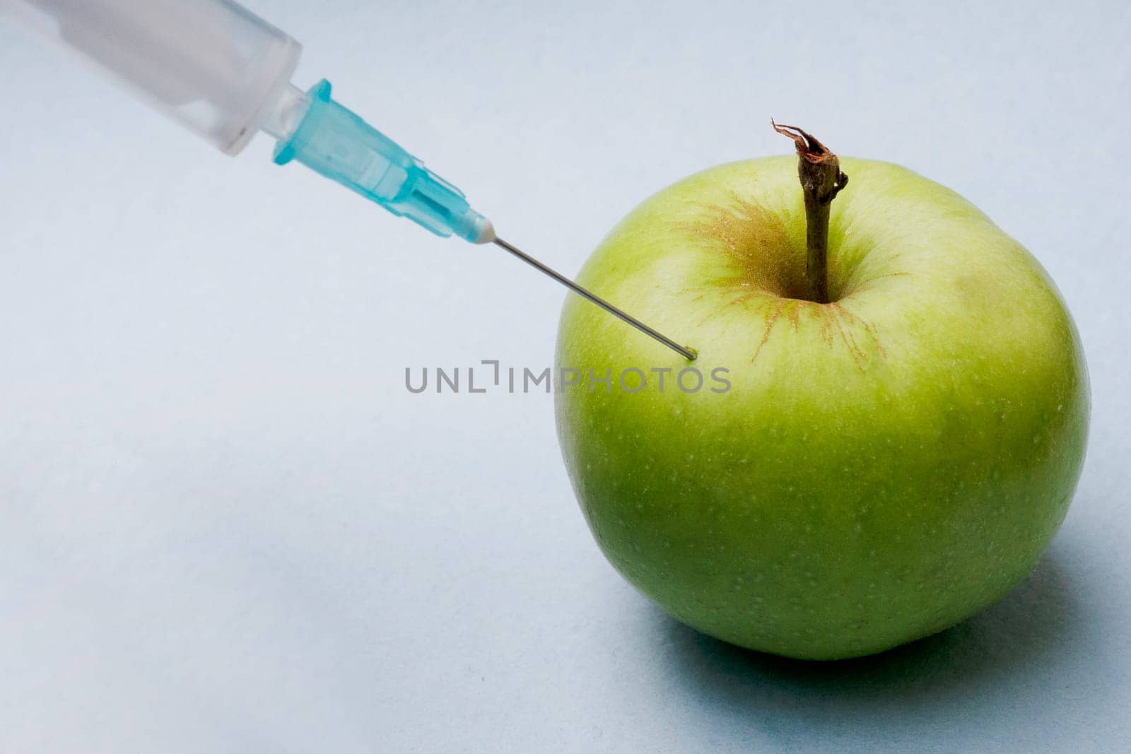 A syringe with a needle pierced a green apple close-up on a blue background. GMO concept photo.