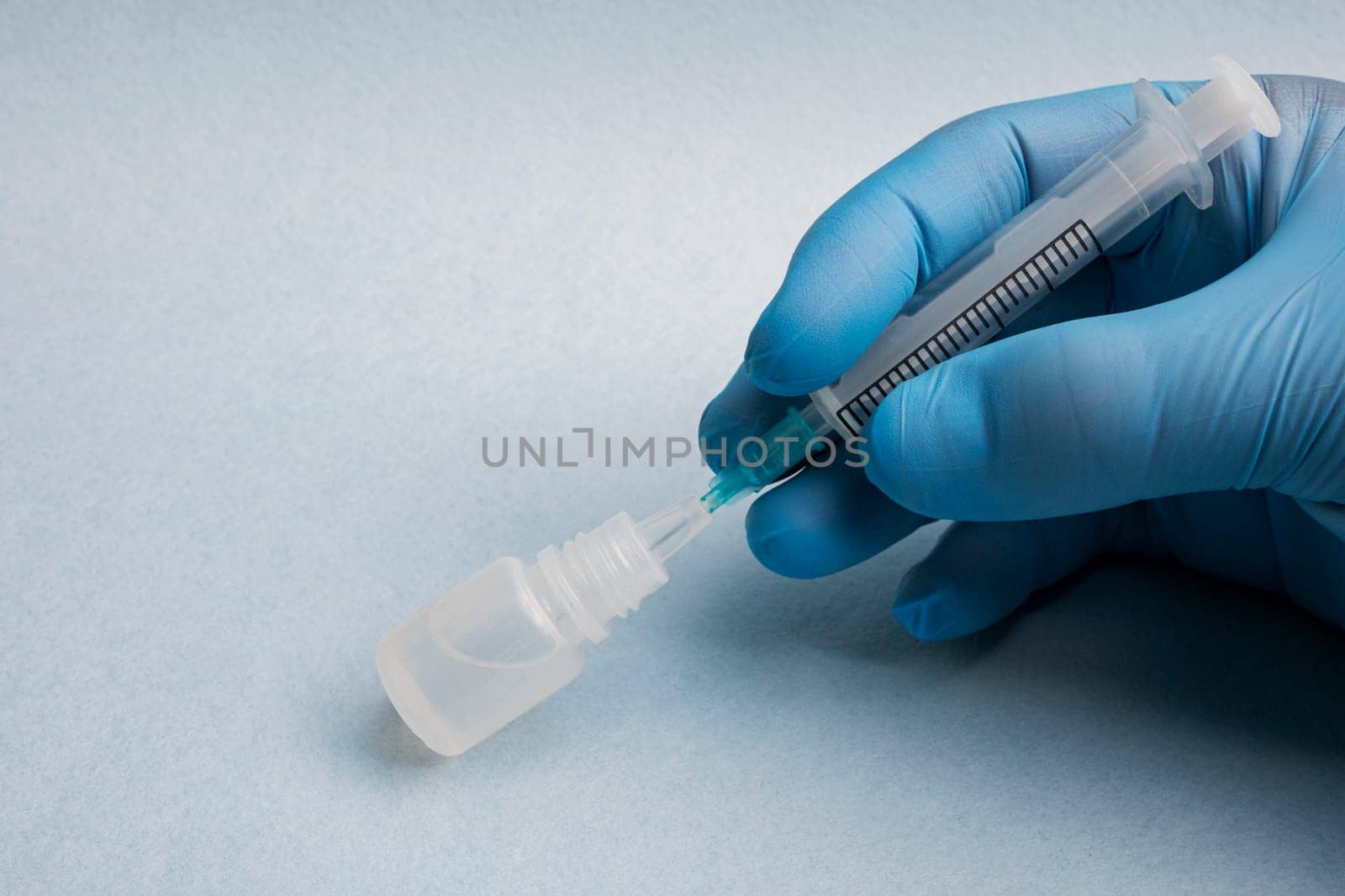 Close-up of a doctor's hand in a blue glove inserting a syringe into a bottle of medicine on a blue background.