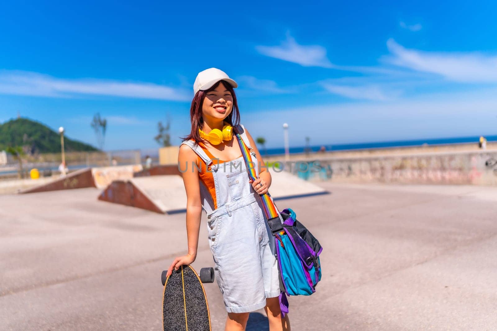 Cute chinese woman in denim clothes and cap standing in a skateboard park in a sunny day holding an skate