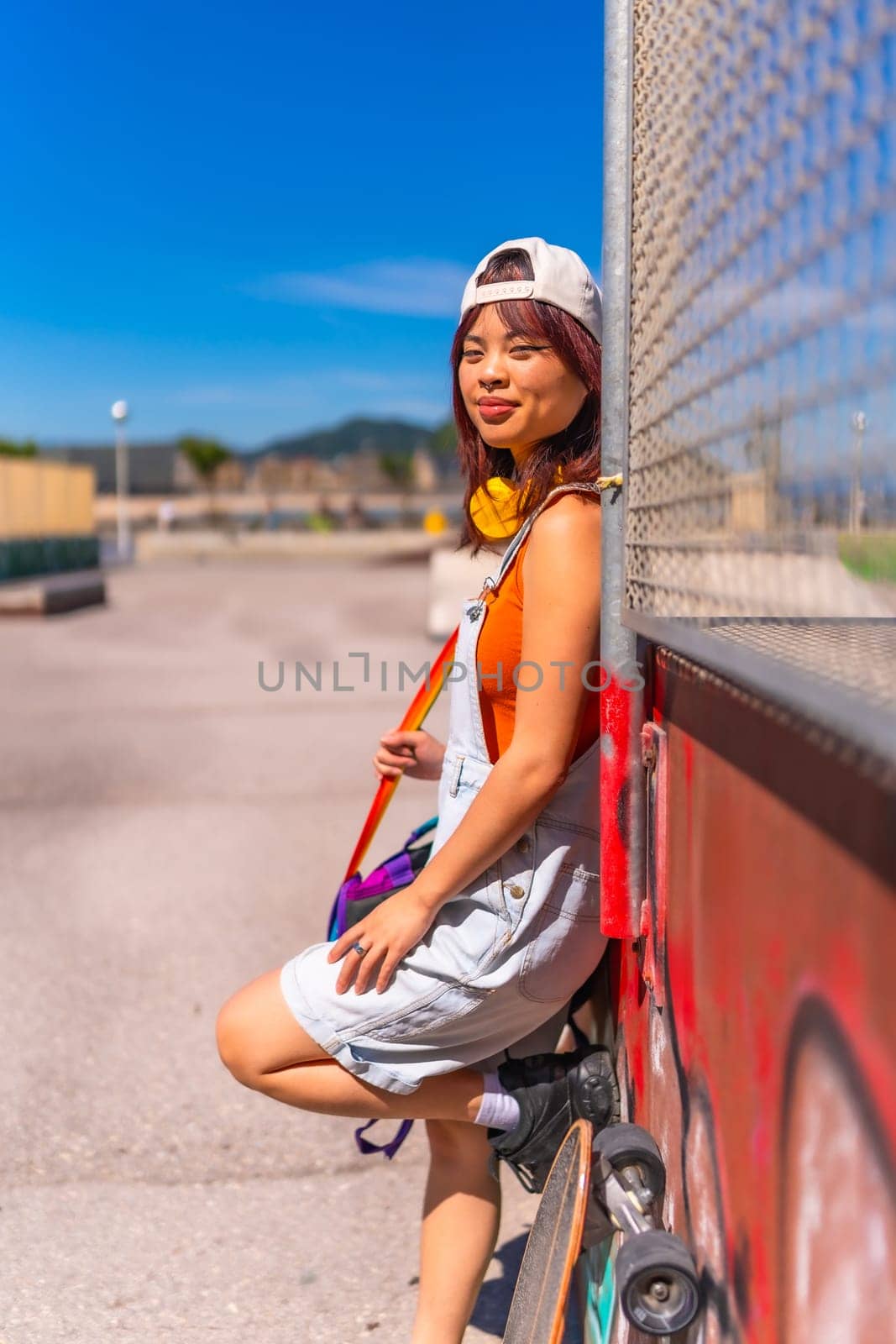 Vertical side view portrait of a young asian female skater leaning on an urban wall smiling at camera