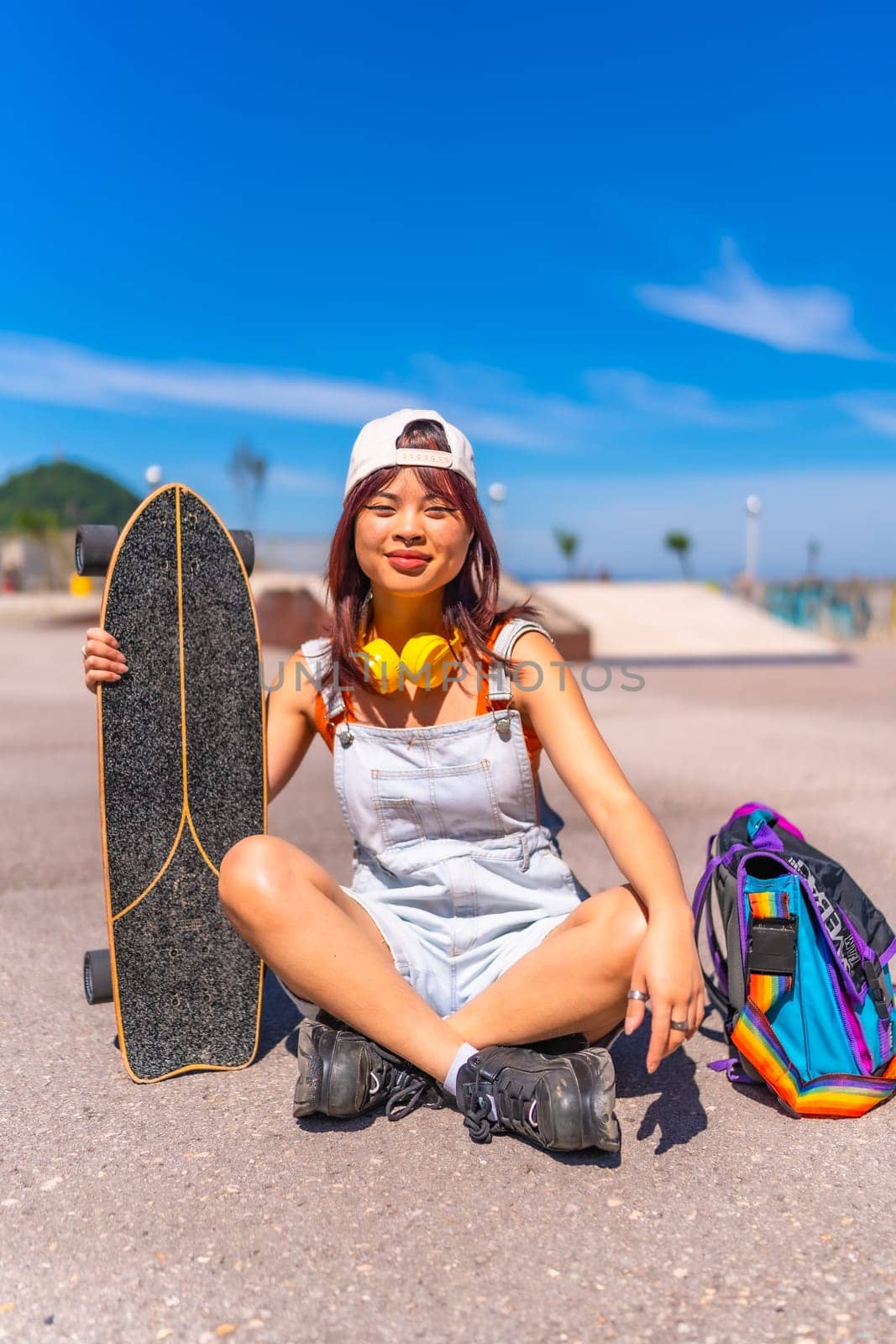 Chinese female skater wearing casual clothes, cap and headphones sitting on a urban park in summer