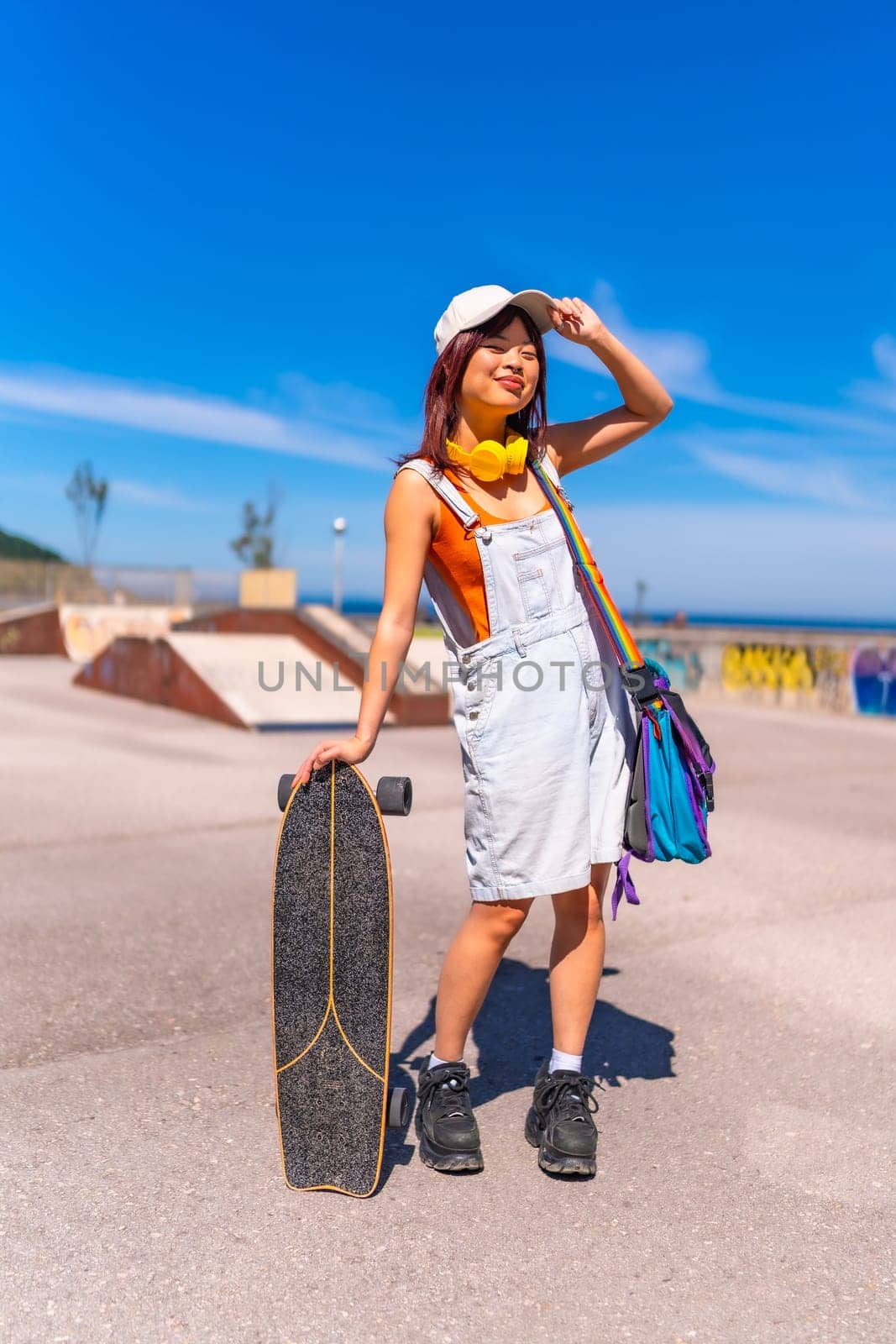 Asian woman holding a skateboard and adjusting her cap standing on a skateboard park