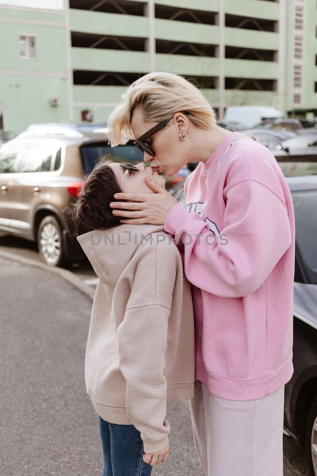 Candid moment between a mother and her daughter standing in a parking lot. Both are dressed in casual, fashionable outfits with sunglasses, emphasizing a relaxed and stylish urban lifestyle.