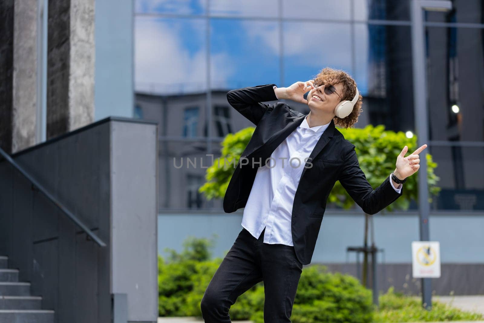 Happy relaxed young Caucasian businessman in wireless headphones dancing, listening favorite energetic disco music dancing outdoors. Overjoyed guy in suit outside office building in downtown city.