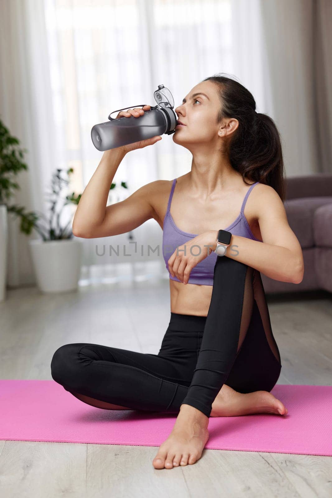 athletic fitness caucasian woman drinking water sitting on pink exercise mat at home