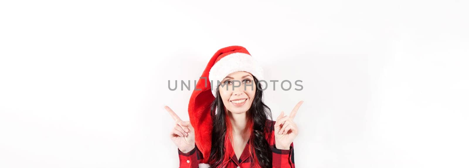 Smiling woman in red Christmas hat holding finger at empty Space for text for promo discount. Beautiful lady, Long curly hair, red santa hat, red pajama. Woman portrait isolated on white background