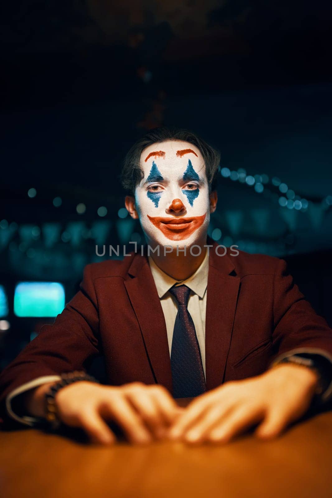 Man in suit and tie with the Joker makeup sits at the table. Halloween party in nightclub with decorations and scary costumes. Autumn holiday. All Saints' Night. Photo card with copy space.