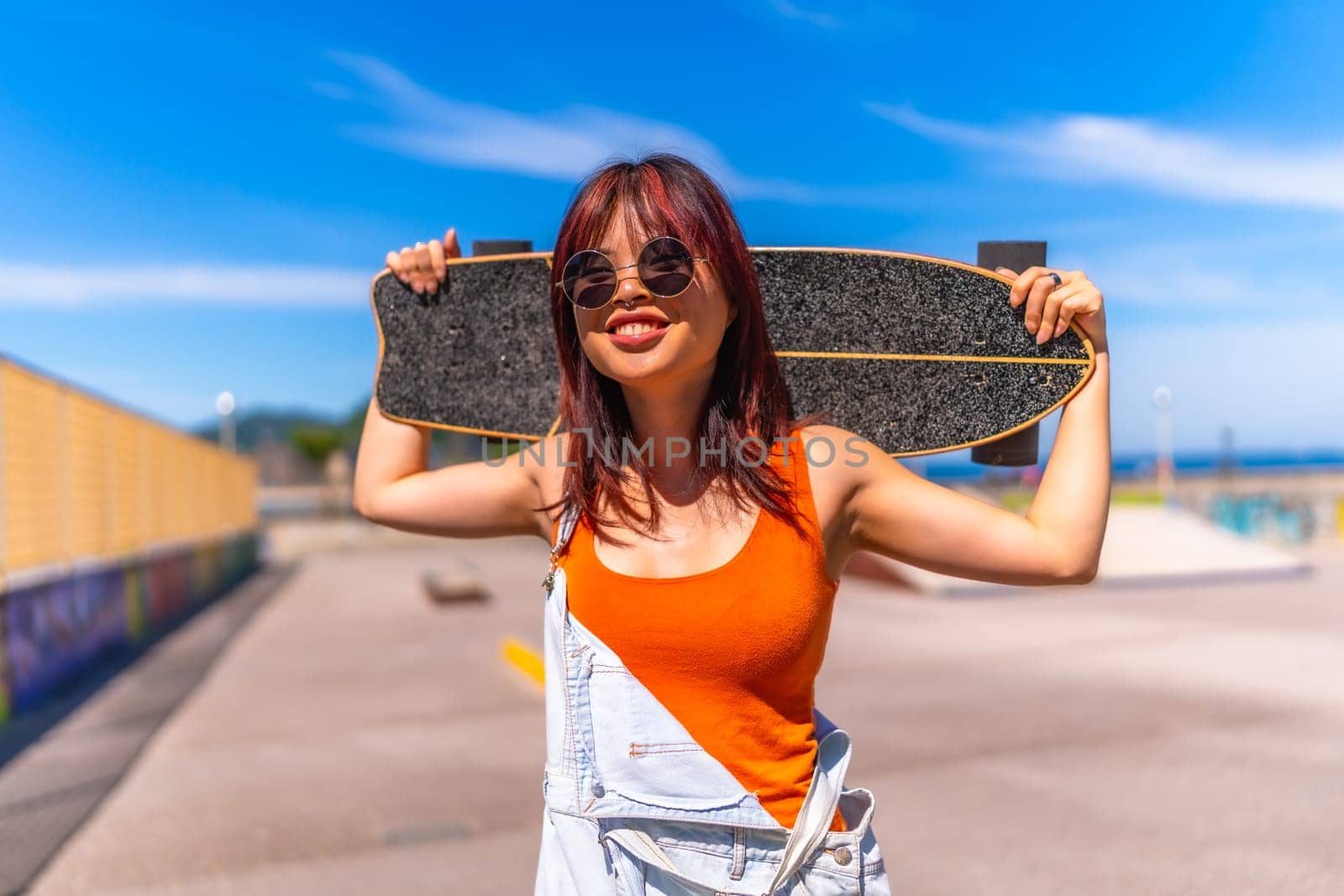 Portrait of a cool woman in dungarees and sunglasses holding on shoulders her skateboard on an urban park