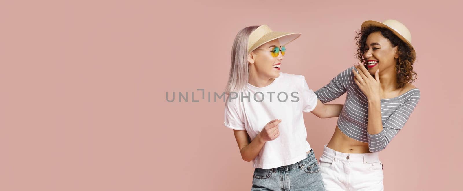 This image shows two women, one with blonde hair and wearing a visor and sunglasses, and the other with dark curly hair, laughing together in front of a pale pink background, copy space