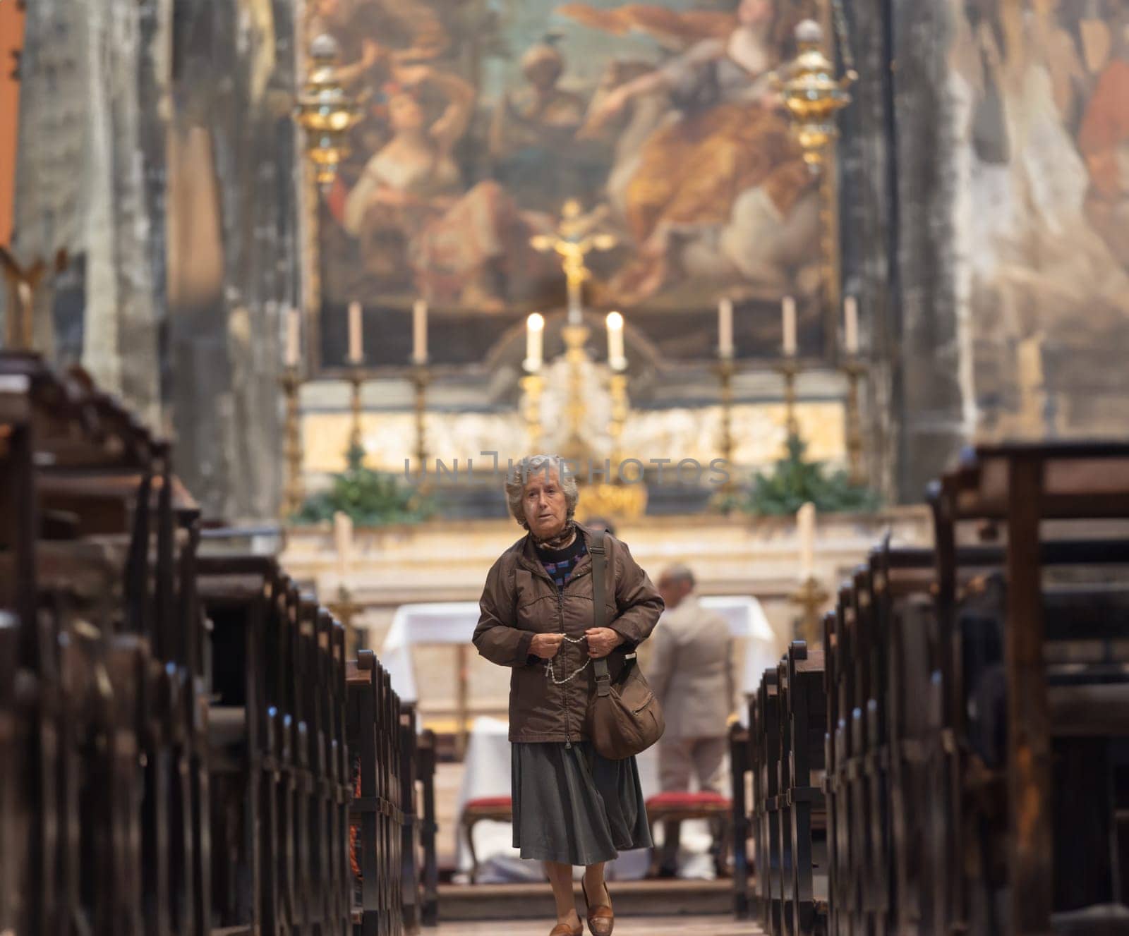 8 december 2023, Lisbon, Portugal - an old woman is walking down a long aisle of a church. She is wearing a brown coat and a scarf