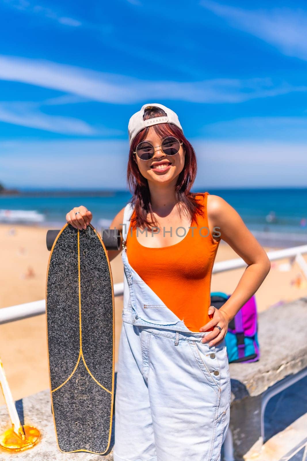 Vertical Portrait of a cool young female asian skater in summer modern clothes, cap and sunglasses smiling at camera standing next to the beach