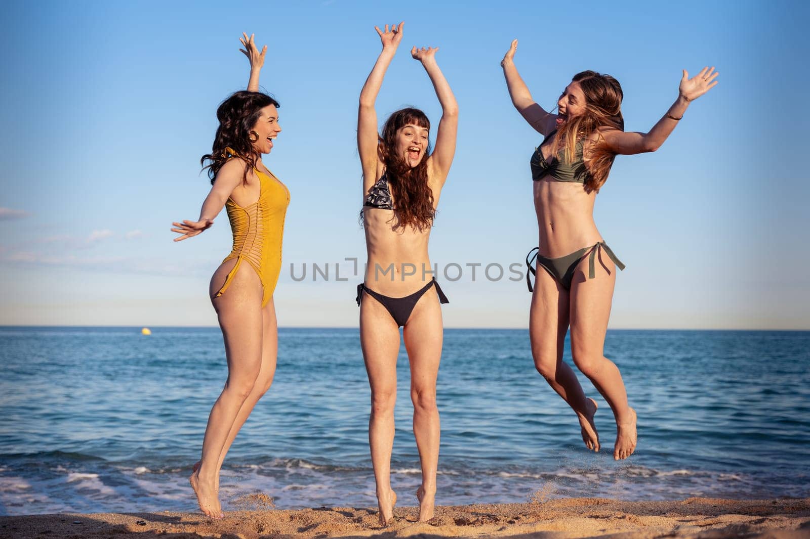 Three happy girls in jumping motion with on the beach outdoors in summer celebrating vacation days. Friends having fun on vacation by the sea carefree, and party concept