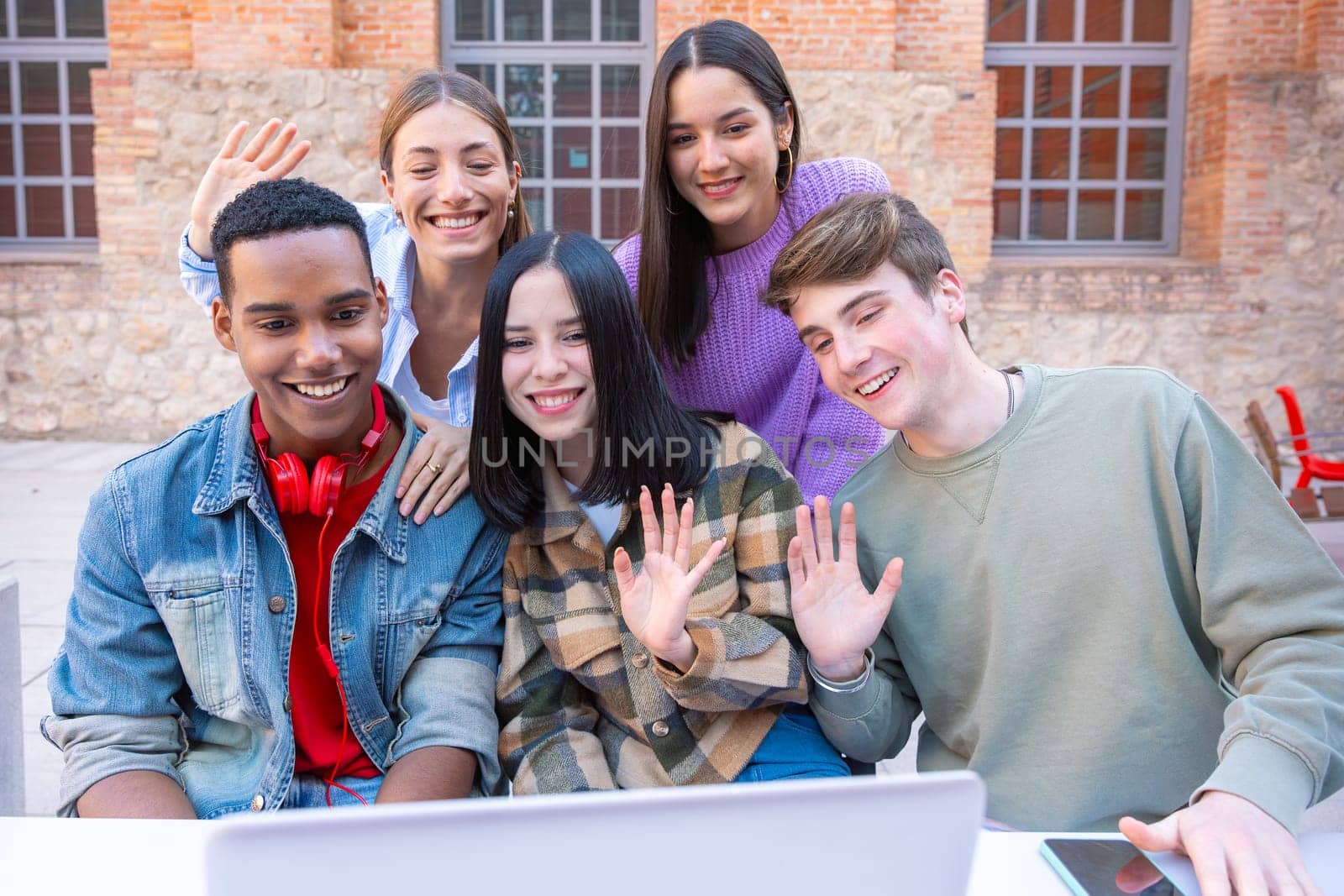 Group of happy, smiling students on the college campus make a video call on a laptop. Five students greet and laugh. Concept: university life