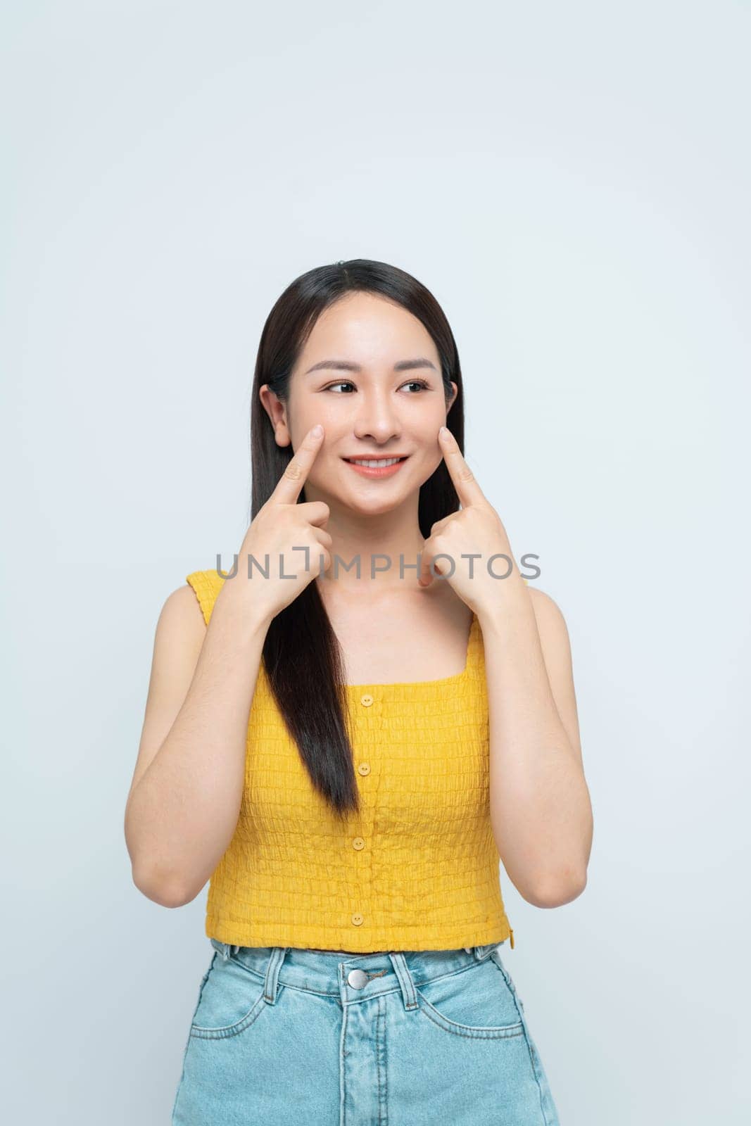 Cheerful woman pointing to her cheeks, expressing happiness and positivity on a light background.
