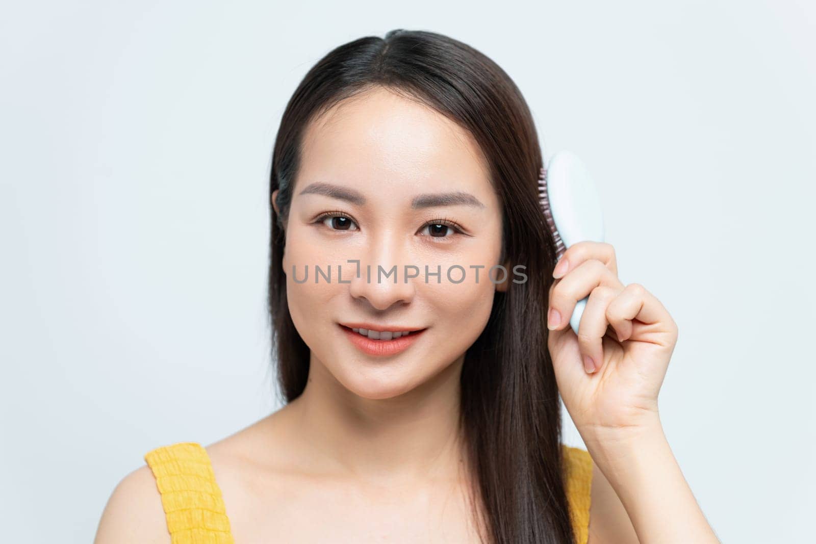 Smiling woman brushing her hair with a white hairbrush, wearing a yellow top and blue jeans.
