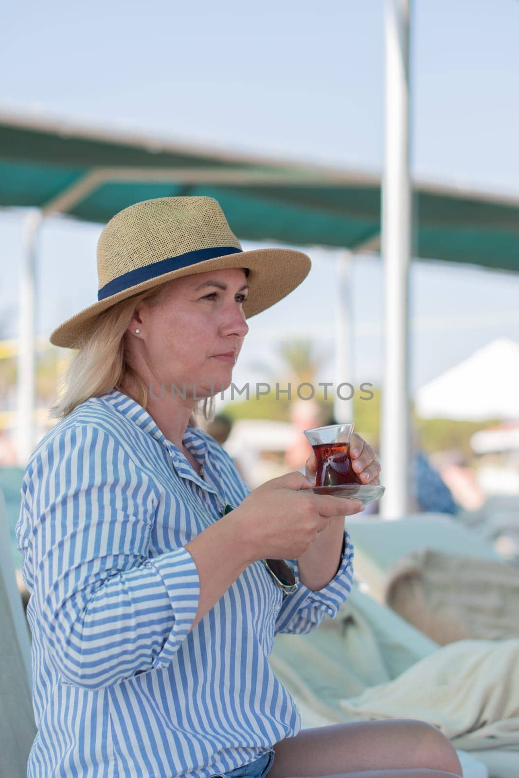 Young blonde drinks tea from Turkish Armudu cup,sitting on sun lounger on the sea beach, and looks at the sea, being in deep thought,wearing a long blue and white striped shirt and a straw hat