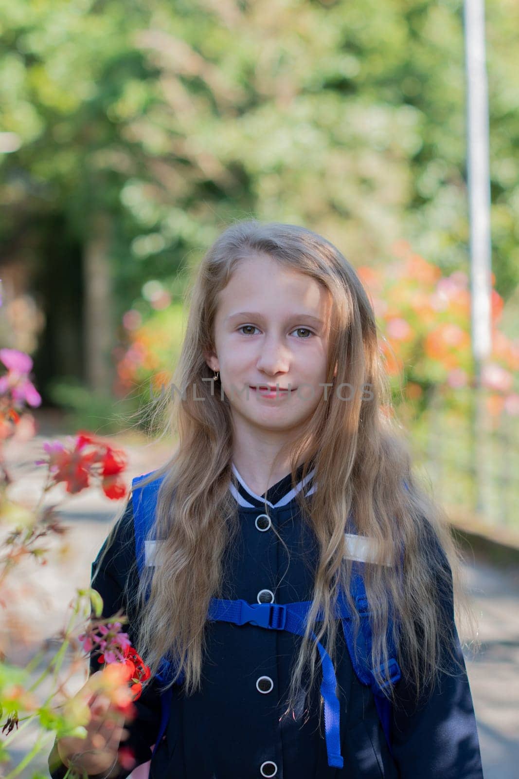 Back to school, portrait of a little girl from primary school outdoors, enjoying flowers in a flowerbed and looking at the camera with a smile, student dressed in dark blue uniform