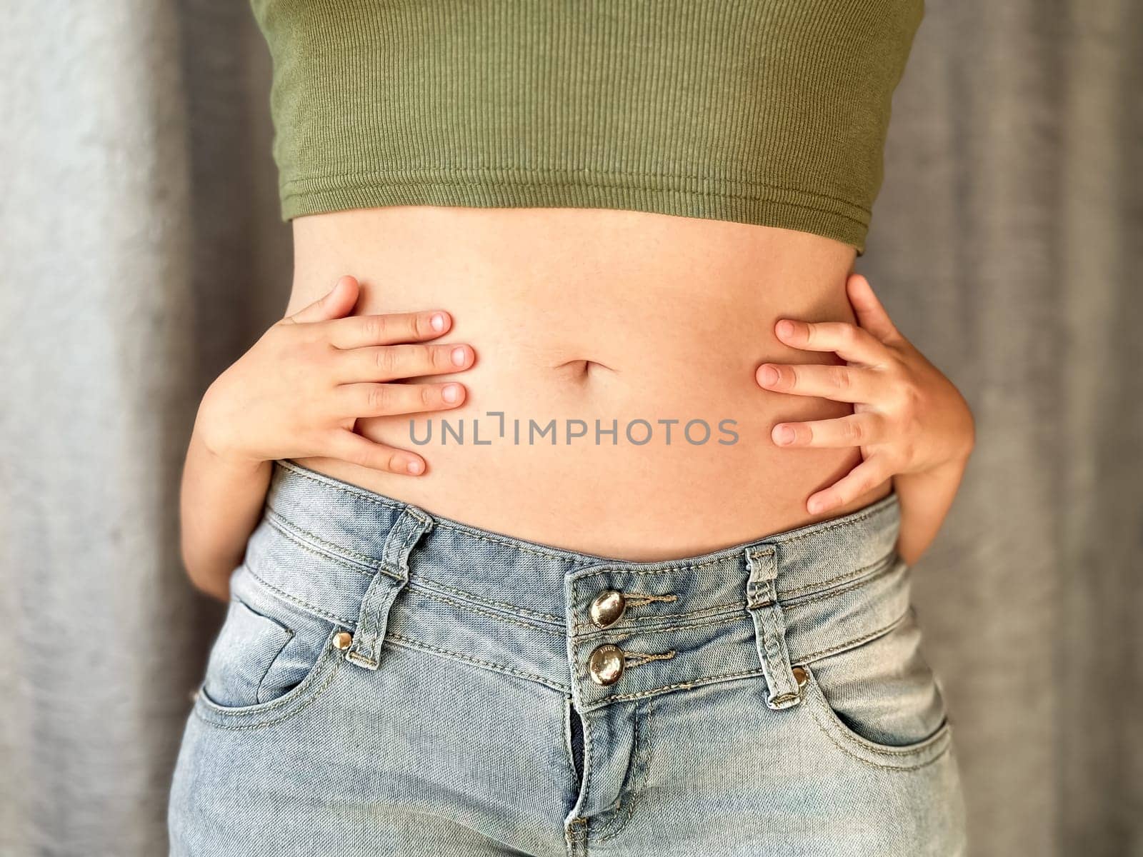 Close up of woman in jeans and green top with childs hands around her waist. Concept of family bonding, motherhood, postpartum recovery and affection. Neutral background. High quality photo