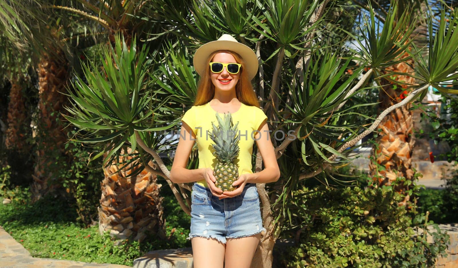 Summer vacation, tourism, happy smiling young woman with fruits, pineapple, joyful girl in glasses, straw tourist hat posing on the beach with palm tree