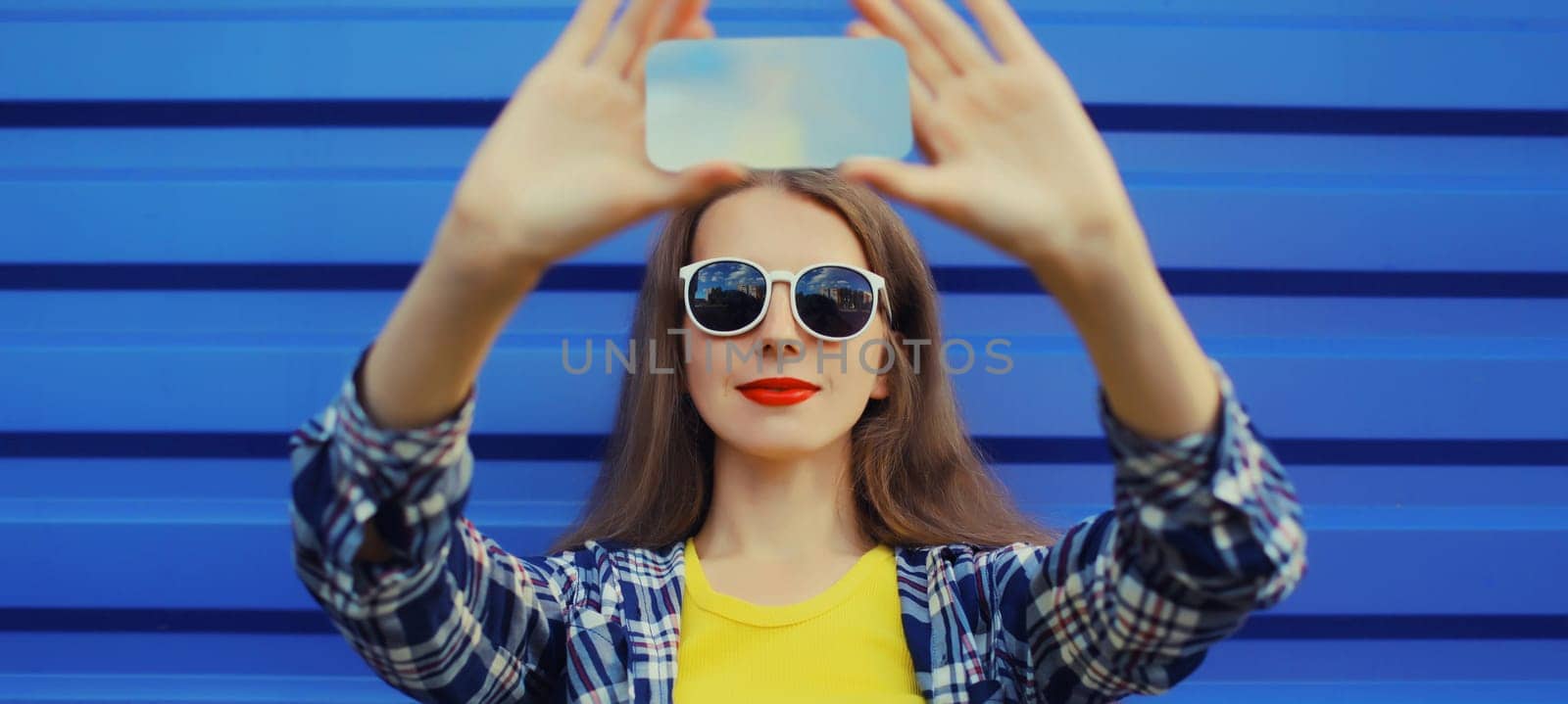 Close up of modern young woman taking selfie with phone, stylish cool girl using smartphone on blue wall background
