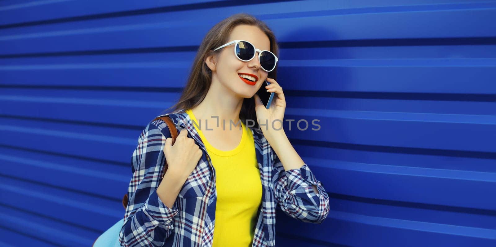 Happy young woman calling on phone, girl talking on the smartphone in glasses looks away on blue wall background