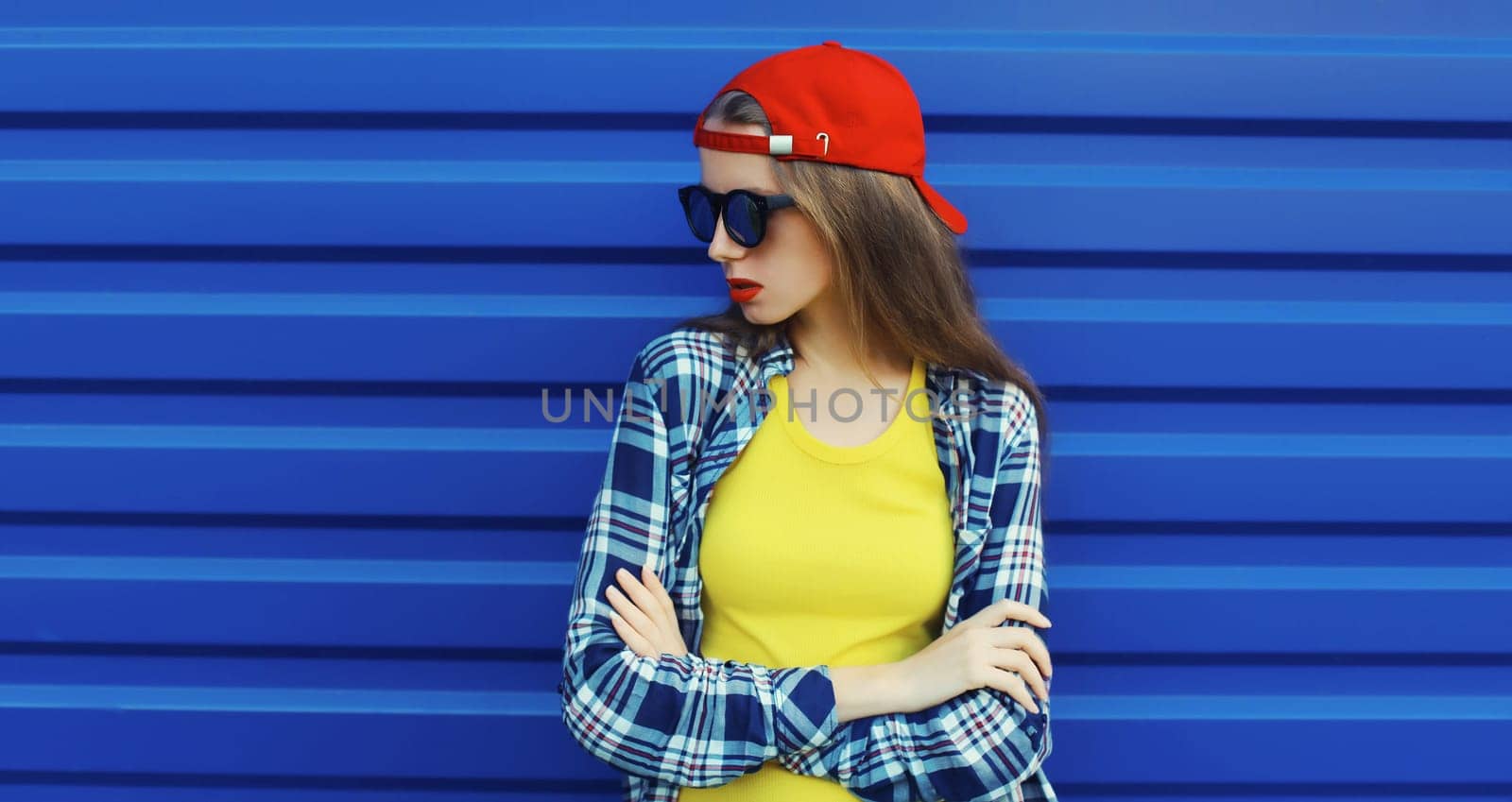 Portrait of stylish young girl in red baseball cap, glasses, modern teenager girl looking away posing on blue wall background