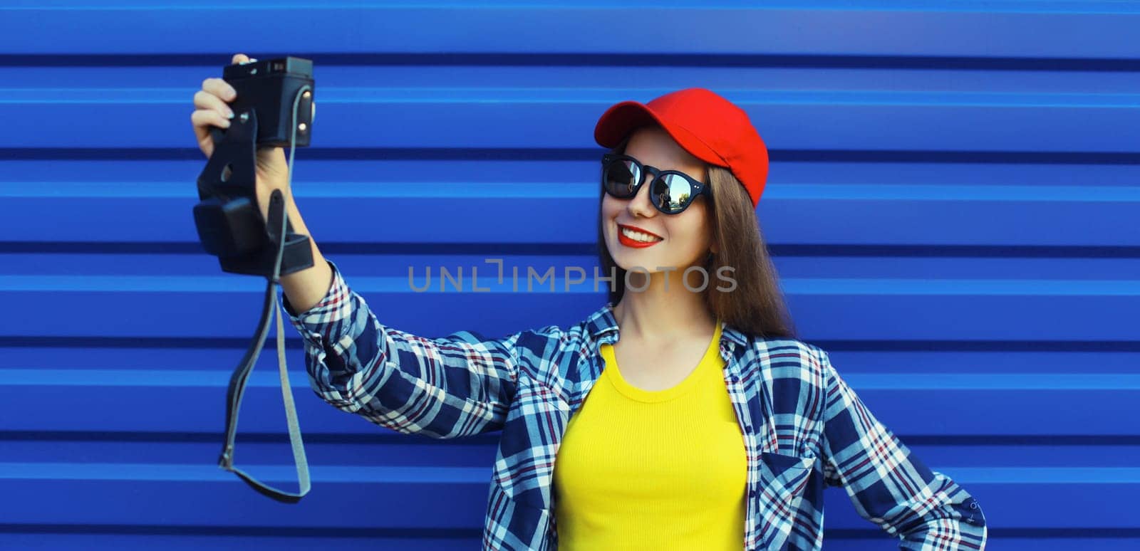 Summer portrait of happy cheerful smiling young woman taking selfie with camera, modern cool girl in red baseball cap on blue wall background