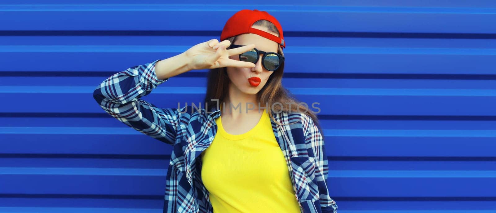 Stylish young woman blowing kiss in glasses, cool modern teenage girl in red baseball cap posing on blue wall background