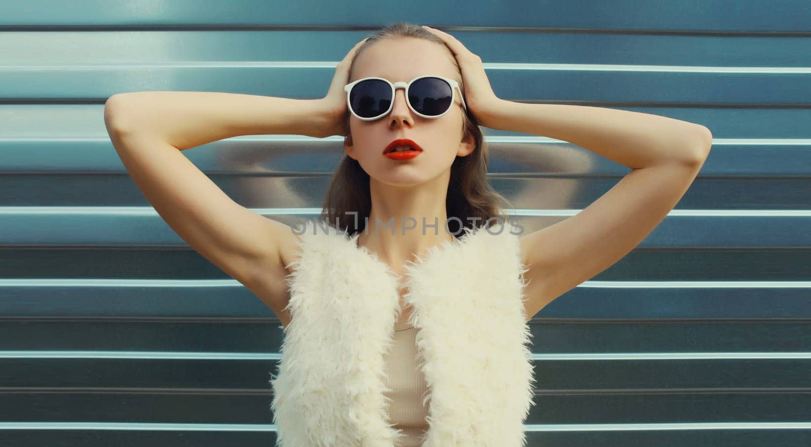Portrait of modern stylish young woman in white fur vest, glasses, fashionable female model posing on gray silver wall background