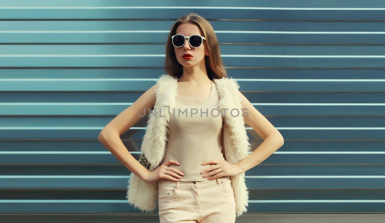 Portrait of modern stylish young woman in white fur vest, glasses, fashionable female model posing on gray silver wall background