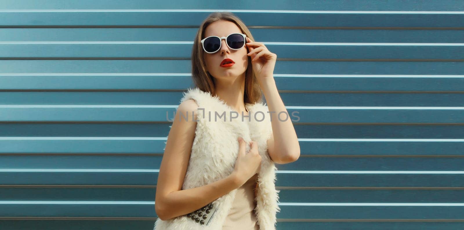 Portrait of modern stylish young woman in white fur vest, glasses, fashionable female model posing on gray silver wall background
