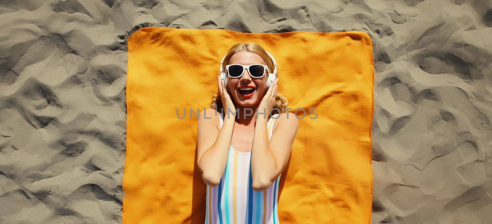 Summer vacation, tourism, happy cheerful young woman listening to music lying on sand on the beach, relaxed tourist girl in headphones on towel in swimsuit, glasses, sunny day, top view
