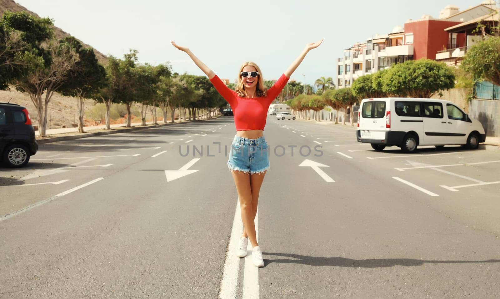 Happy cheerful caucasian young woman raising her hands up standing full-length on the road on a summer day