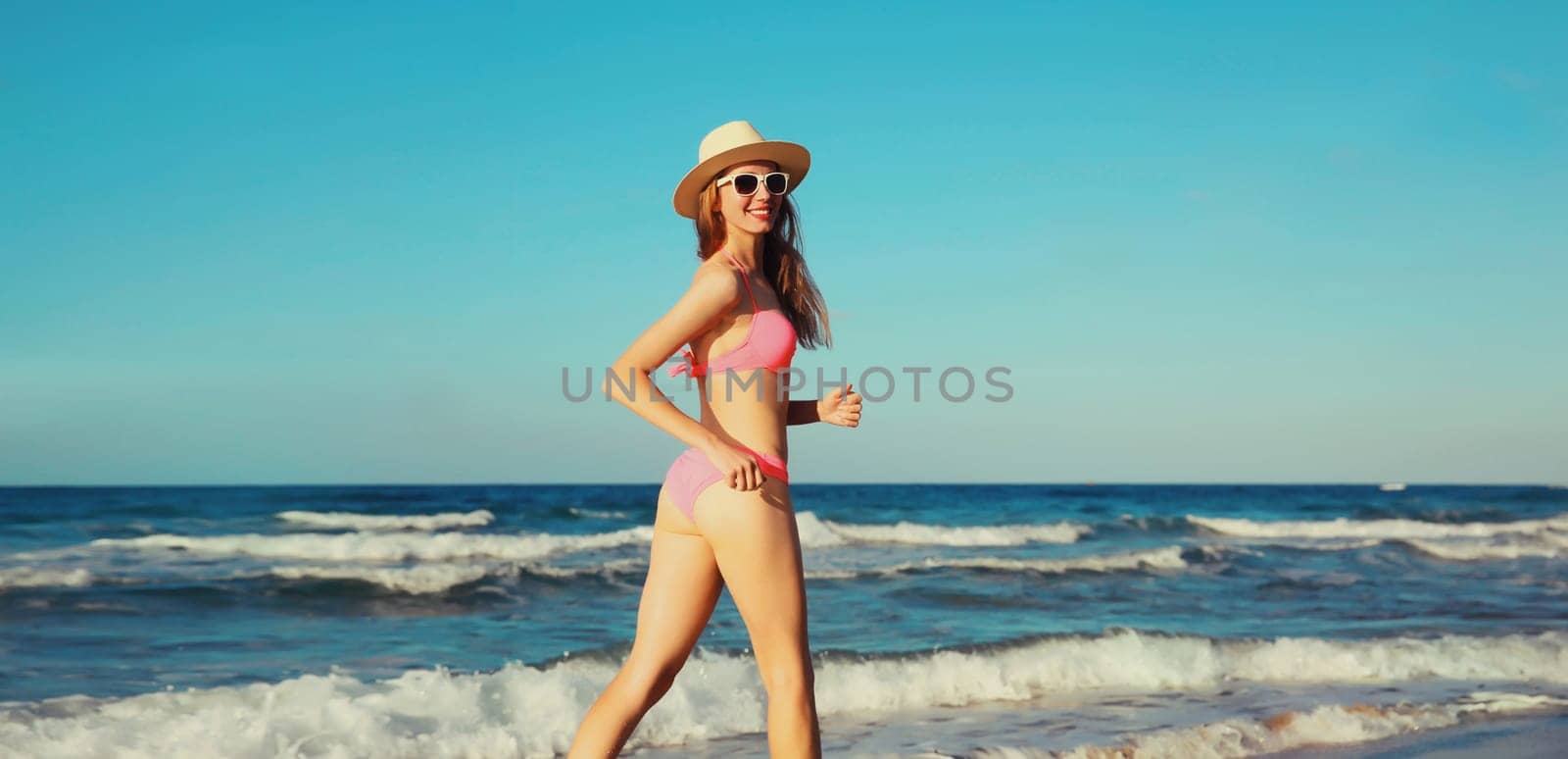 Summer vacation, happy cheerful woman walking running in bikini swimsuit and tourist straw hat on the beach on sea coast with waves background on sunny day