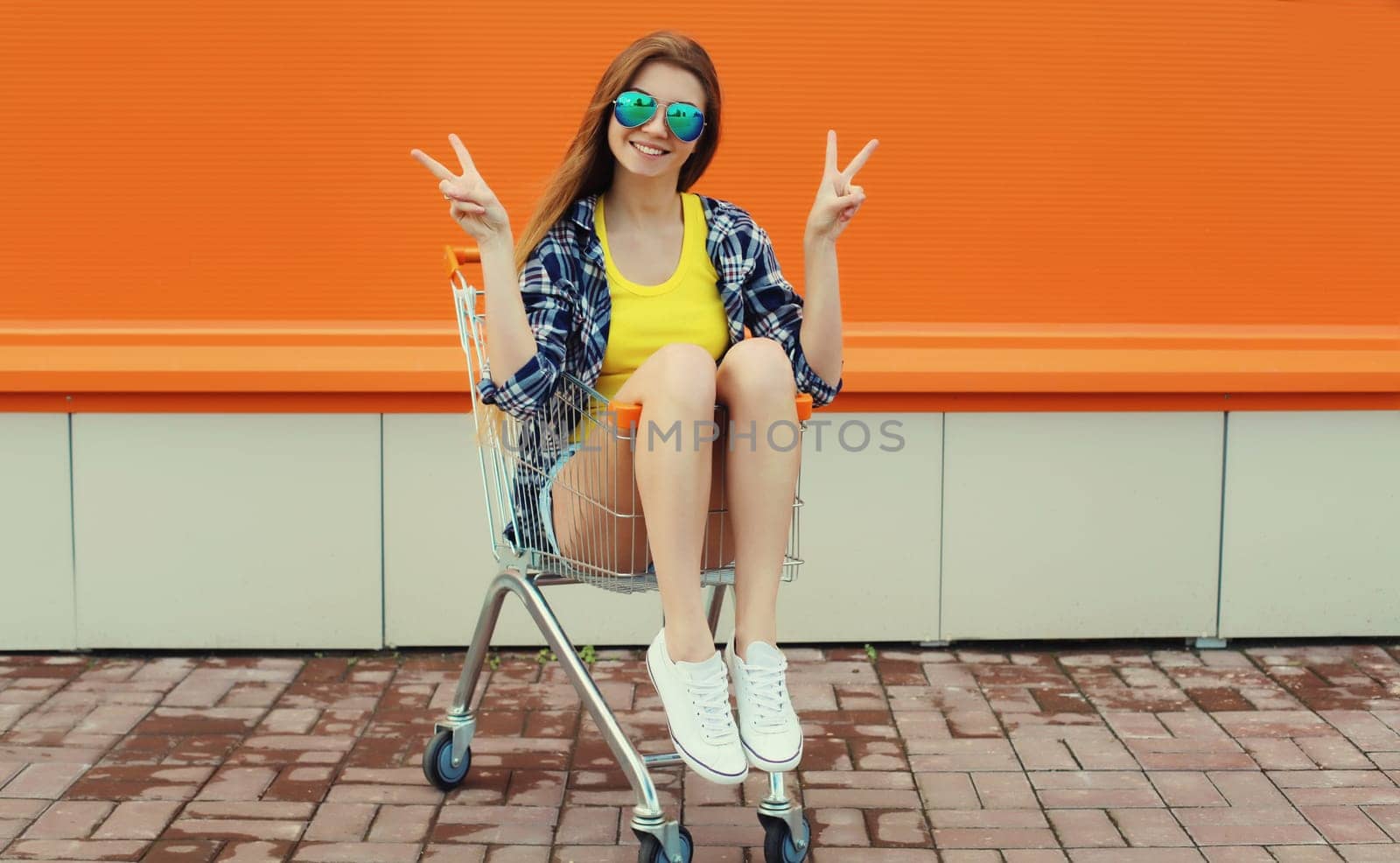 Stylish happy cheerful young woman, modern teenage girl smiling having fun sitting in shopping trolley cart on colorful orange background on city street