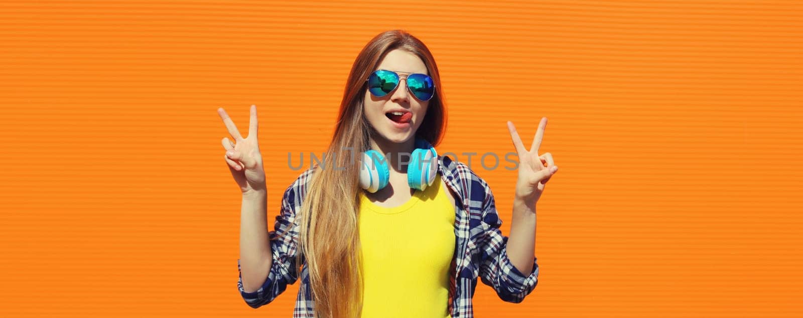 Stylish young woman listening to music with headphones, cool modern teenager girl posing on colorful bright orange background, summer day