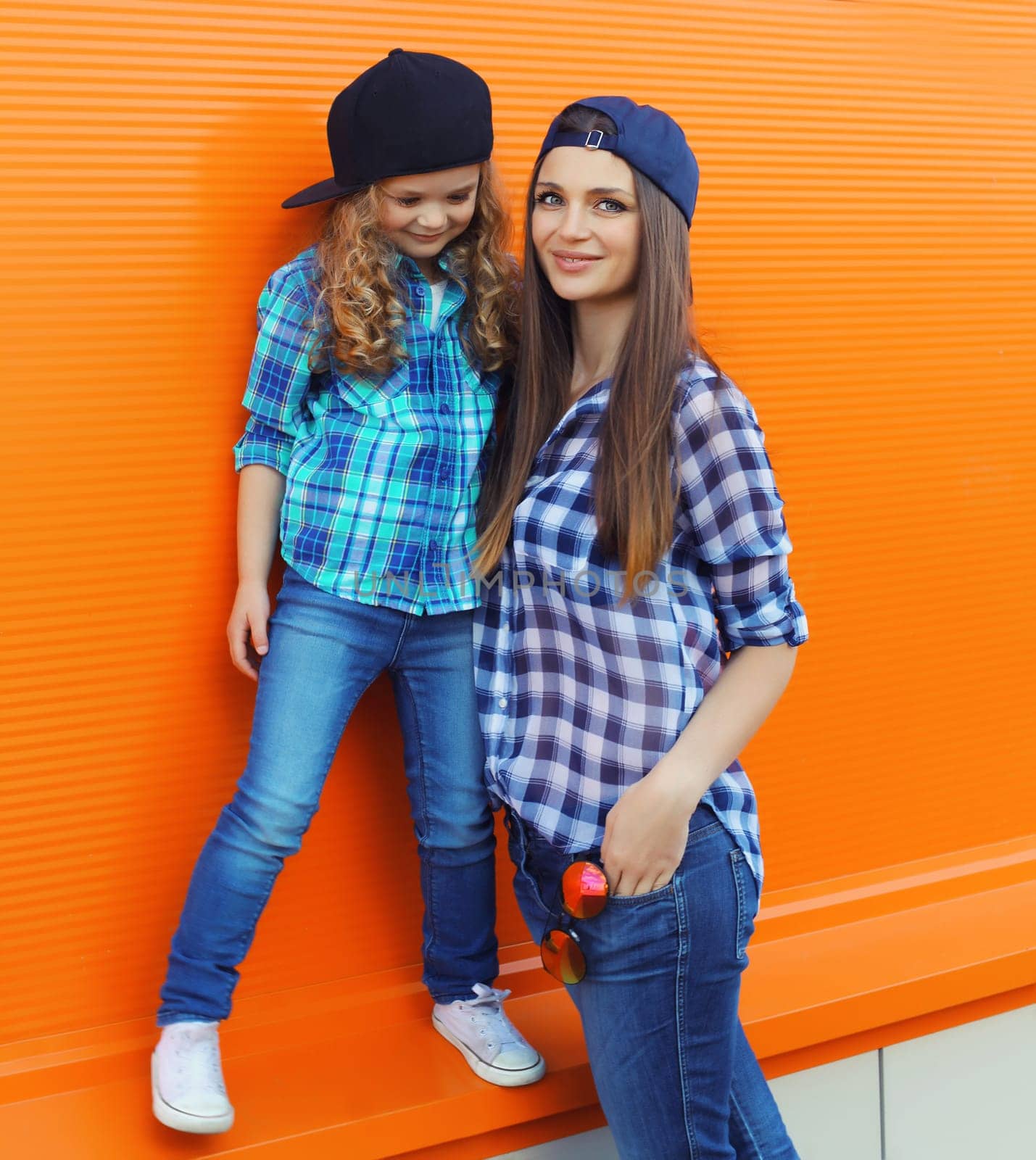 Beautiful stylish mother and child daughter together, happy mom with kid girl in baseball cap, shirt, casual clothing in the city on orange background