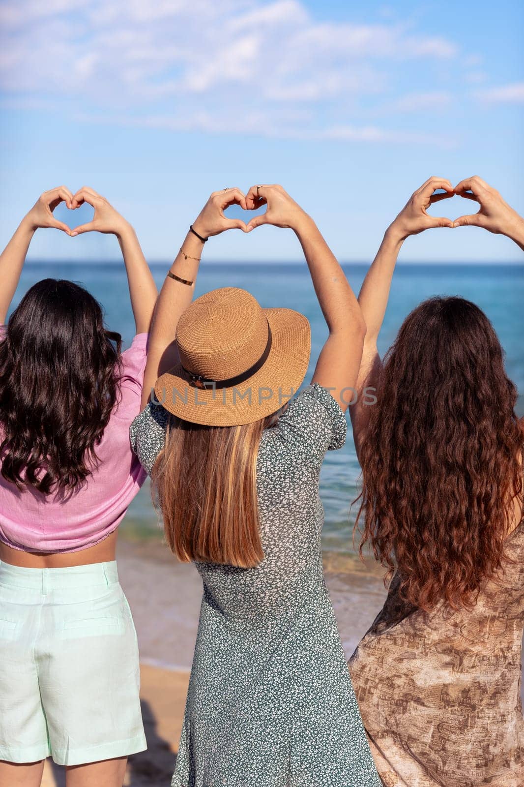 Rear view three young female friends on vacation on the beach making heart shapes with hands shot from behind with sea background. vertical