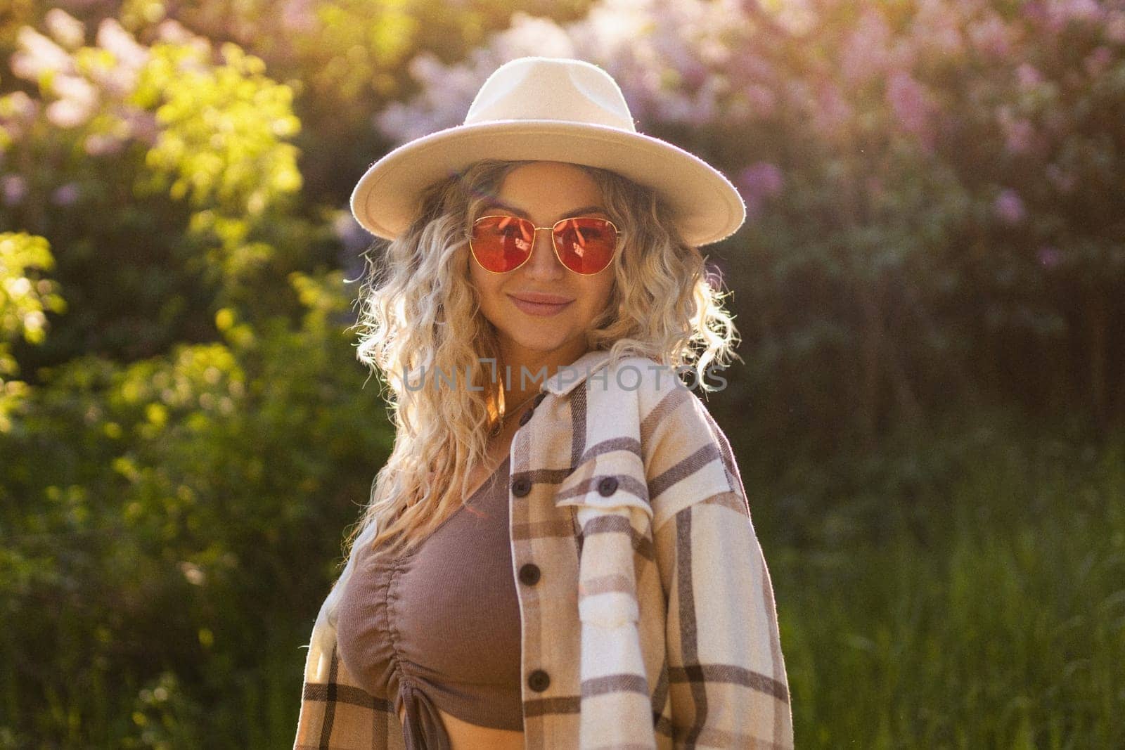 sexy curly blonde in a hat and sunglasses posing near blooming trees.