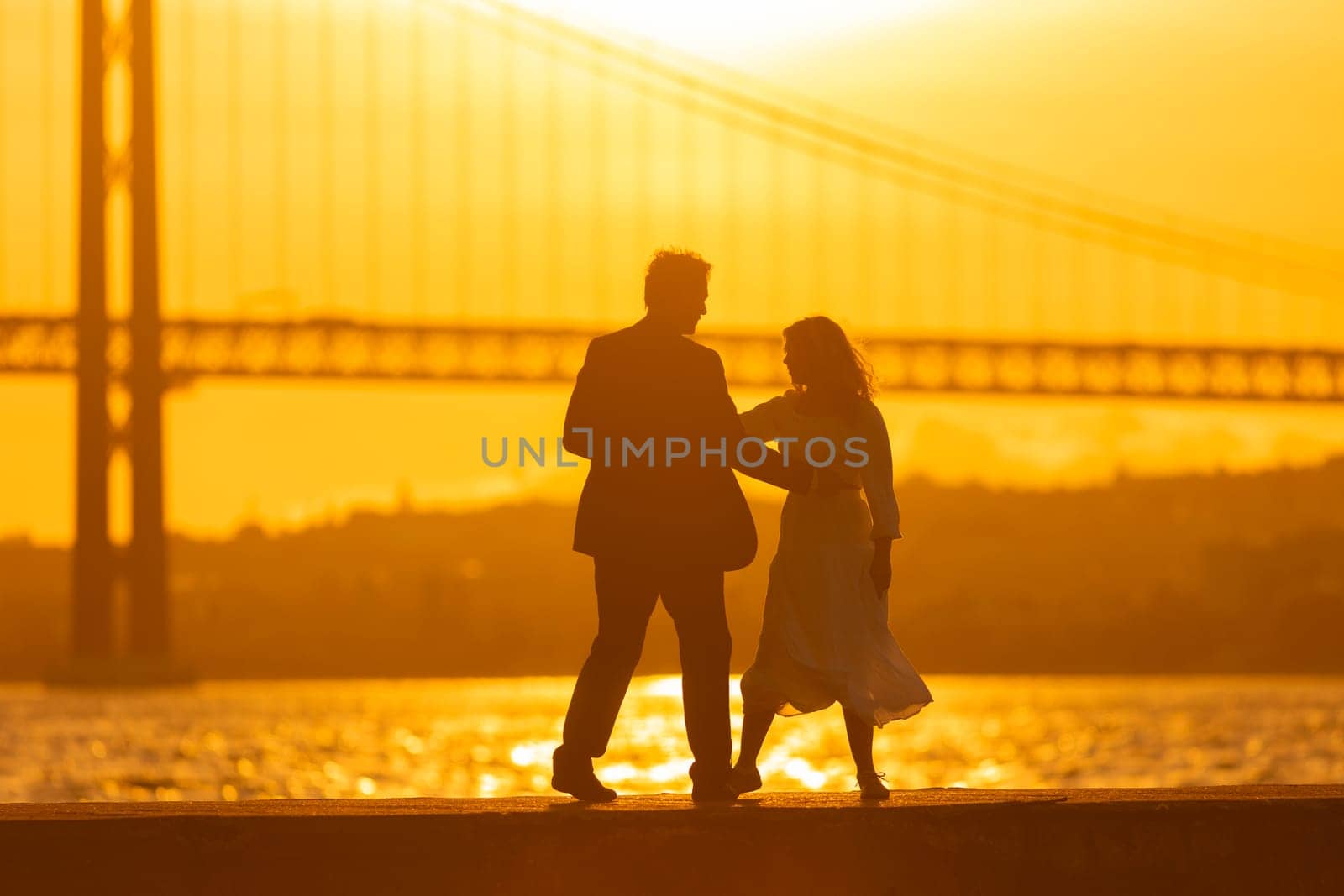 A couple dances on a dock in front of a bridge at sunset. The golden light of the setting sun creates a silhouette of the couple.