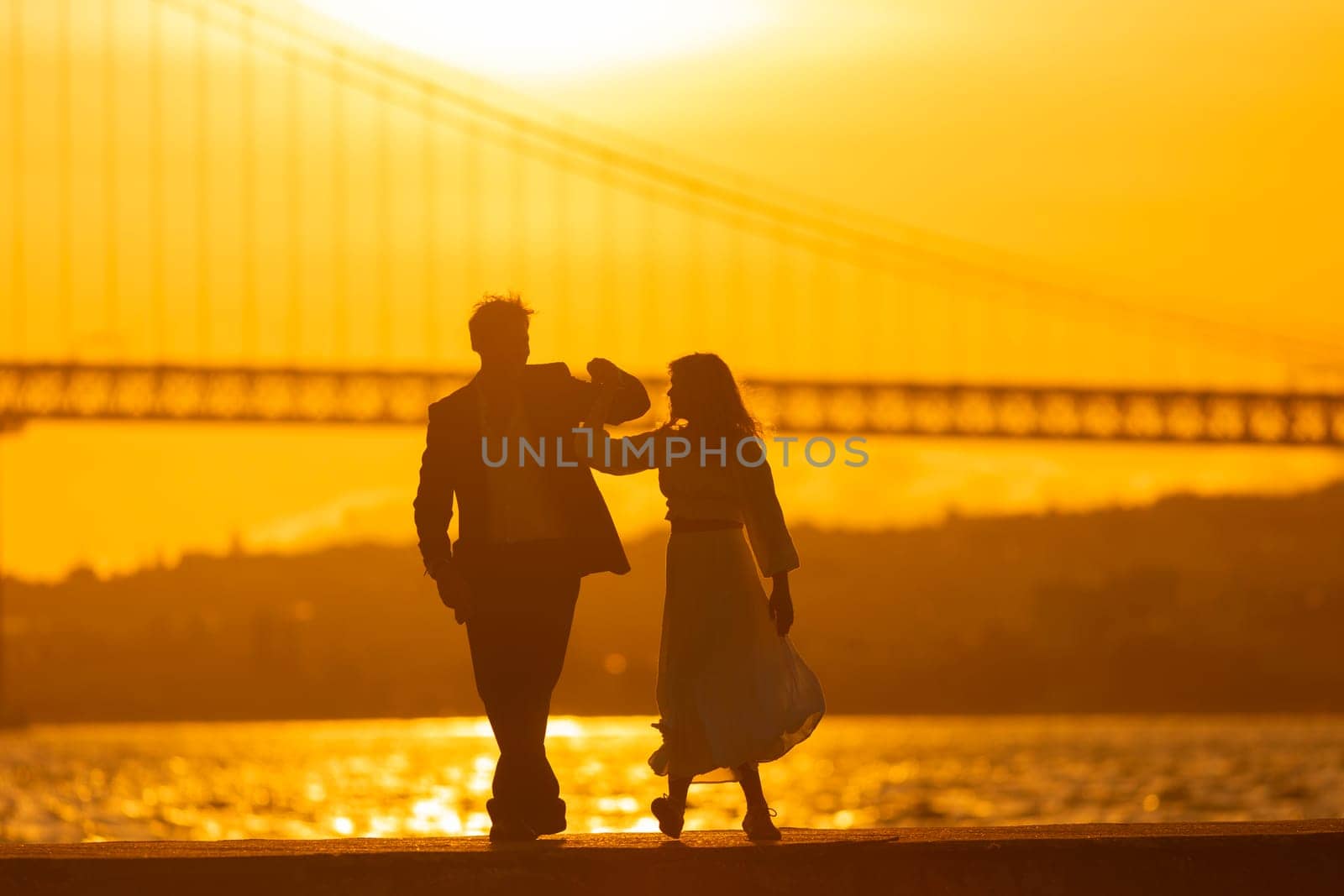 A man and woman dance in silhouette against a sunset with a bridge in the background.