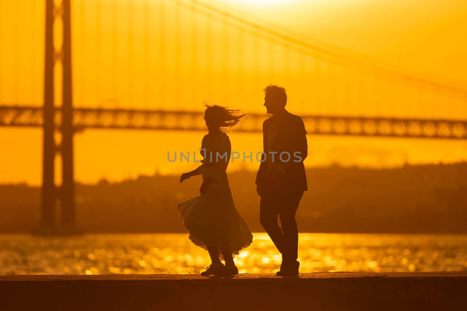 A couple dances silhouetted against a sunset with a bridge in the background.