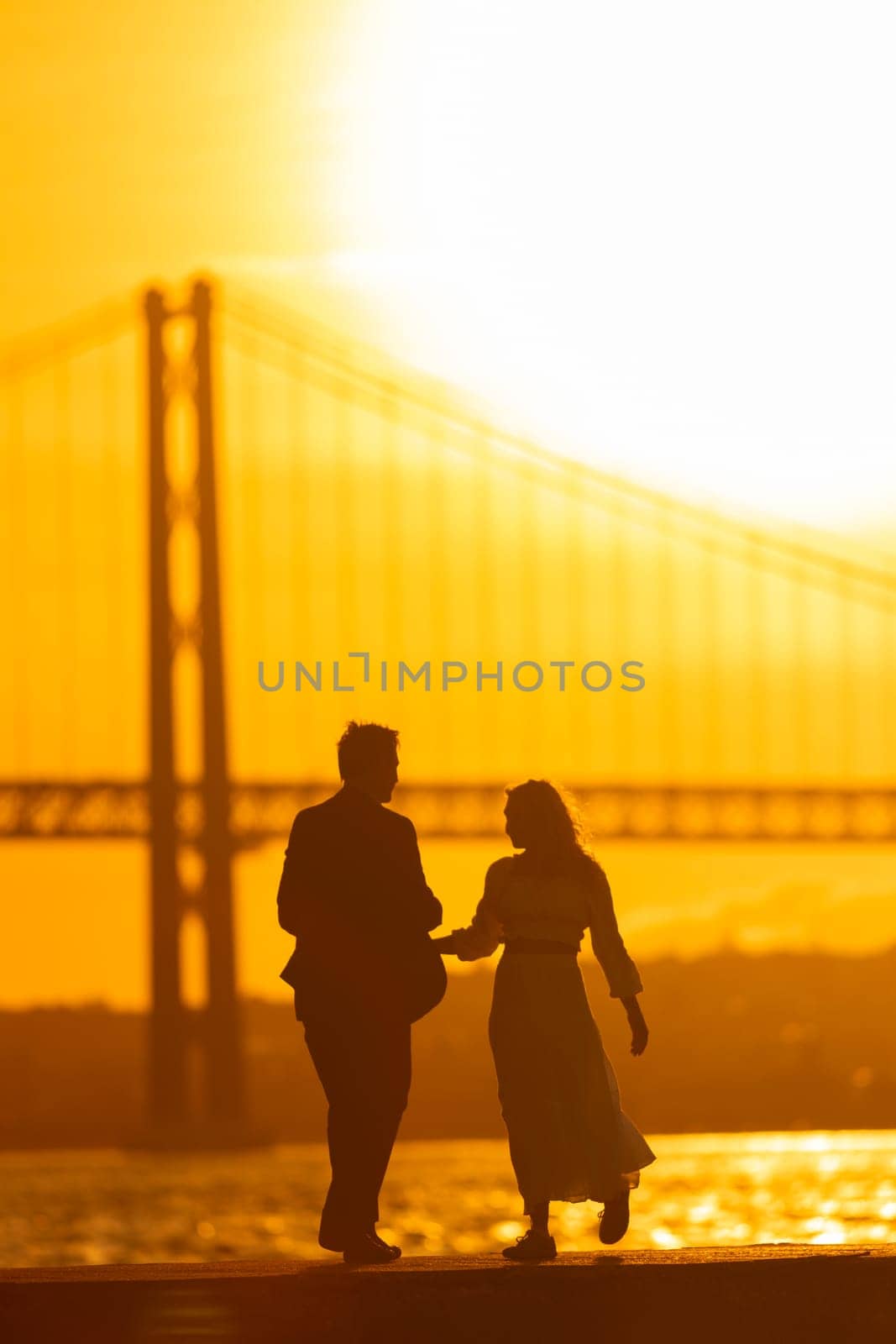 A couple dances silhouetted against a vibrant sunset, with the iconic 25 de Abril Bridge in the background.